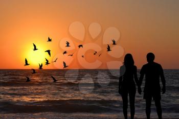 View of a full body of couple silhouettes holding hands looking at sunrise on the beach