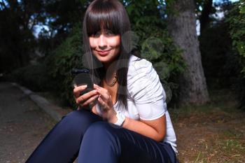 Smiling teenage girl shows cell phone, against green of summer park in the dark closeup