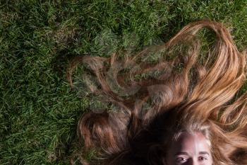 Long blond beautiful hair is spread over summer grass. We can see only half of the face - only pretty eyes.  Beautiful young girl is lying on green grass in the evening time.