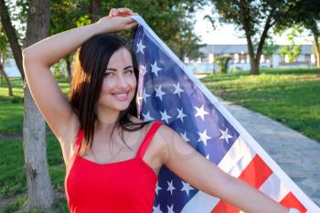 Happy young woman holding american flag in her hands. She weared in pretty red dress.