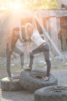 Brunette and blonde posing standing on giant tires outdoors at sunset backlit by sun
