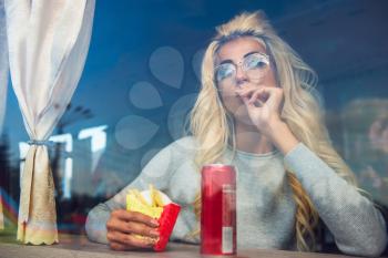 Fast food and leisure concept - beauty young blonde woman eating a fried potato in the cafe