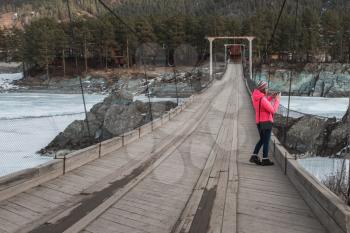 Happy woman taking photos on the bridge from river at beauty sunny spring day, in Altai mountains. Travel vacation concept.
