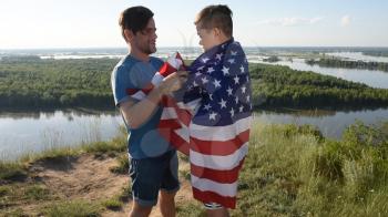 Father with American flag embraces his son on river bank. Patriotism, independence day 4th july concept