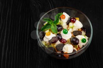 Salad from grapes apples pears kiwi oranges with mascarpone chease and cream. Healthy fresh fruit summer salad in glass bowl on black wooden background with copy space.