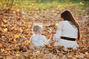 A young mother and little blonde daughter are sitting on fallen leaves in an autumn park, and mom gives her a bouquet of leaves. Mom walks with her daughter in the autumn park.