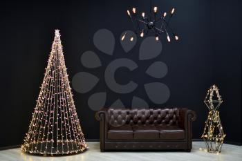 New year's Loft-style decor against a black wall, a Christmas tree from a garland. black chandelier with incandescent lamps and brown sofa. Beautiful New Year's decor with lighting in the studio.