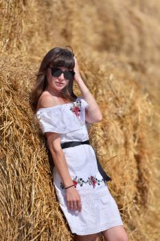 A brunette girl in a white dress and glasses, against a background of a large haystack from round bales.