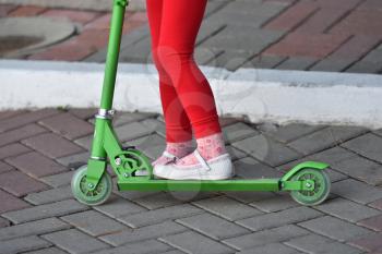 A little girl in red pants and white sandals rides a scooter.