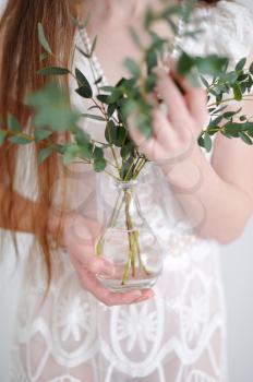The hands of a young girl hold eucalyptus branches that are inserted into a glass vase that is transparent to a small vase.