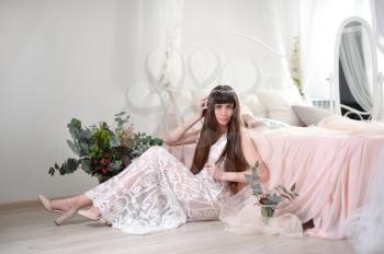 Photo of a young girl in the style of fayn art. A girl in a white translucent dress sits on the floor next to the bed and a bouquet of flowers.