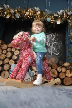 Little girl child sitting on a red rocking horse on the background of the Christmas interior with a fireplace, firewood and gifts