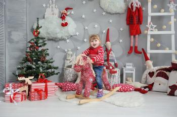 Little girl child sitting on a red rocking horse and smiling on the background of the Christmas interior with a gnomes, gifts and a Christmas tree