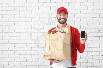 Delivery man holding paper bag with food and phone on brick wall background�