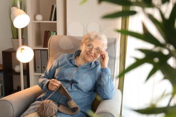 Senior woman sitting on armchair while knitting sweater at home�