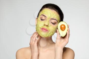 Beautiful young woman with facial mask and fresh avocado on light background�