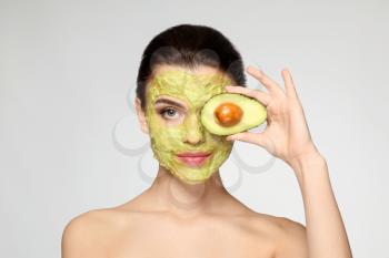 Beautiful young woman with facial mask and fresh avocado on light background�