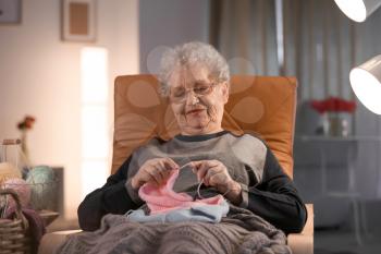 Senior woman knitting warm sweater in armchair at home�