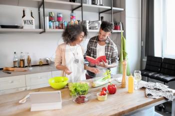 Young African-American couple reading recipe book while cooking together in kitchen�