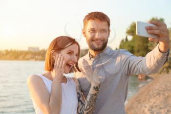 Happy couple taking selfie with engagement ring near river on sunny day�