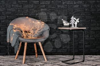 Table with Christmas decorations and stylish armchair near dark brick wall indoors�