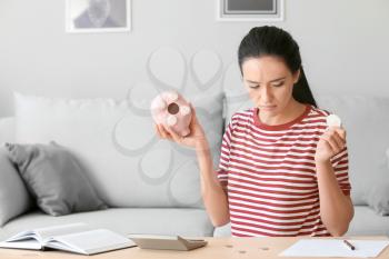 Upset young woman with empty piggy bank at home�