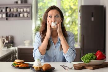 Stressful woman with taped mouth and different products in kitchen. Choice between healthy and unhealthy food�