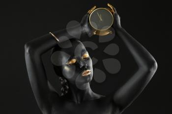 Beautiful woman with black and golden paint on her body holding clock against dark background�