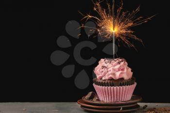 Delicious birthday cupcake with firework candle on table against dark background�