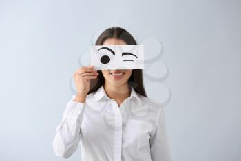 Emotional young woman hiding face behind sheet of paper with drawn eyes on light background�