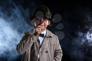 Cute little detective with magnifying glass in smoke on dark background�
