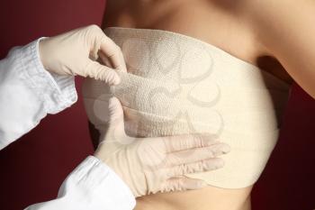 Doctor applying bandage on female chest after cosmetic surgery operation against color background�