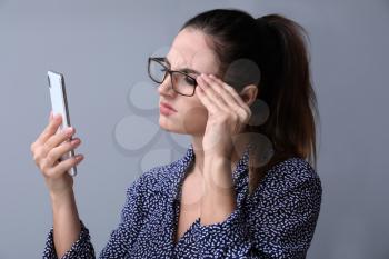 Woman with bad sight trying to read message on screen of mobile phone against grey background�