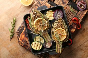 Pan with tasty grilled artichokes and vegetables on wooden table�