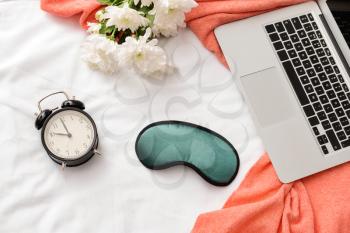 Composition with sleep mask, laptop, clock and flowers on bed�