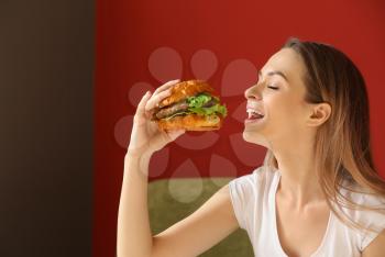 Beautiful young woman eating tasty burger indoors�