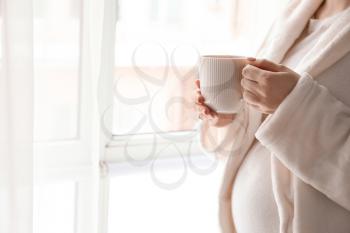 Pregnant woman with cup of hot tea standing near window at home�