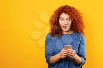 Surprised redhead woman with mobile phone on color background�