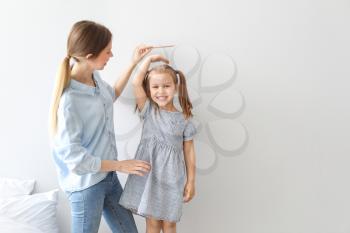 Young mother measuring height of her little daughter near light wall�
