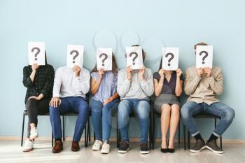 Young people hiding faces behind paper sheets with question marks while waiting for job interview indoors�