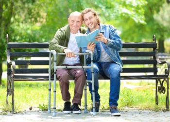 Young man reading book to his elderly father in park�