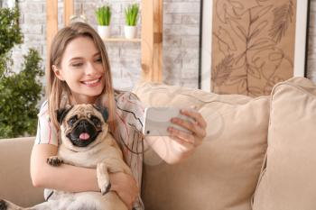 Beautiful young woman with cute pug dog taking selfie at home�