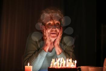 Lonely senior woman with birthday cake in dark room�