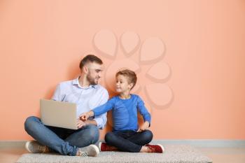 Father and his little son with laptop sitting near color wall�