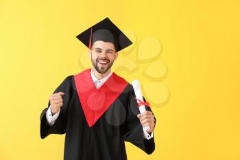Young male student in bachelor robe and with diploma on color background�