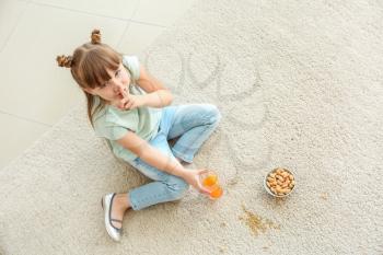 Careless little girl eating nuts and drinking juice while sitting on carpet�