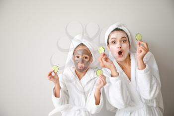 Funny mother and her little daughter with facial masks and cucumber slices on light background�