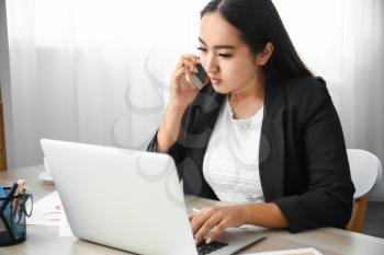 Beautiful Asian businesswoman with laptop talking by mobile phone in office�