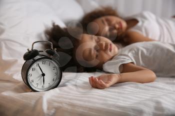 Sleeping African-American girl with mother and alarm clock on bed at night�