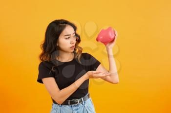 Sad Asian woman with empty piggy bank on color background�
